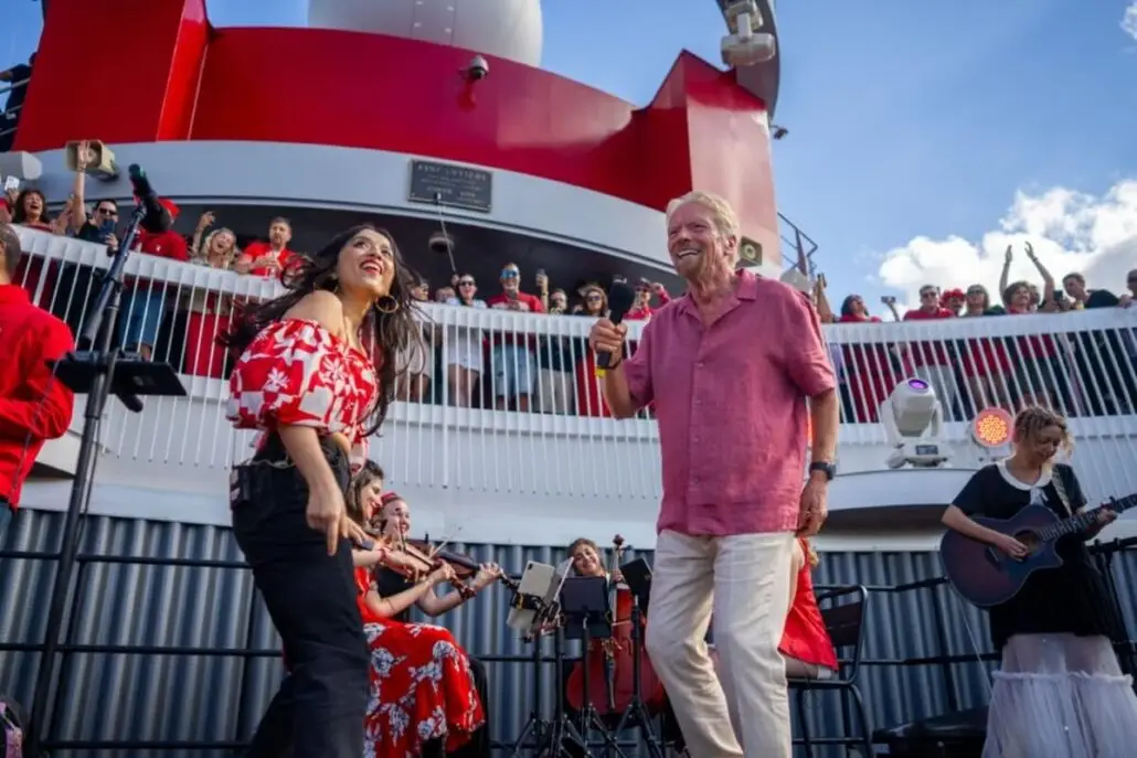 A man in a pink shirt and a woman in a red floral top dance on the Brilliant Lady cruise ship deck near New York, while musicians play string instruments and an audience, possibly including Richard Branson, watches from above.