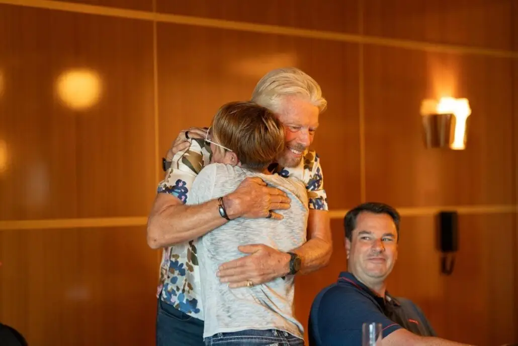 In a warmly lit New York room with wooden walls, a man with gray hair hugs a person wearing glasses—a truly brilliant lady—while another man sits at a table nearby, watching them.