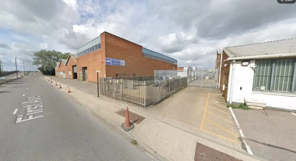A large brick industrial building with a metal fence and gate, possibly a parking facility, sits beside a street labeled "First Ave." Traffic cones line the sidewalk, and a small white building is visible on the right. The sky is mostly cloudy.