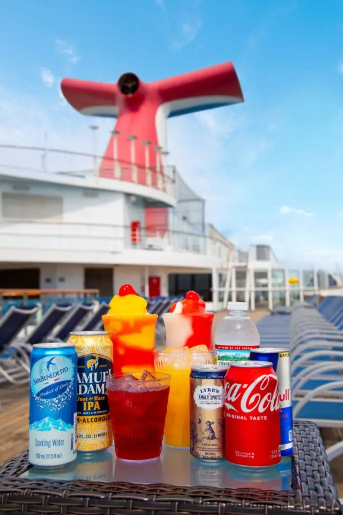 A selection of drinks, including CHEERS! Zero Proof options from the Non-Alcoholic Drink Package, sits on a wicker table on a Carnival cruise ship deck with lounge chairs and a red and white smokestack in the background.