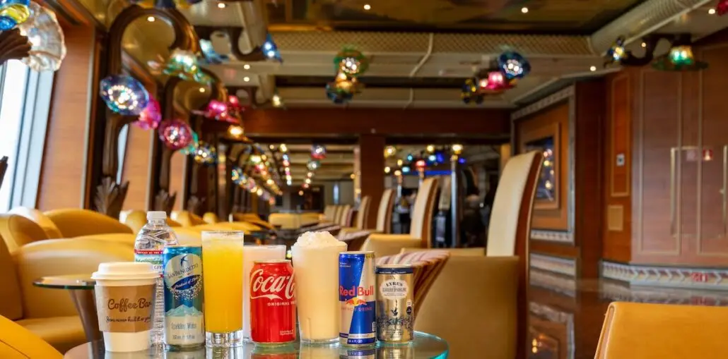 A variety of drinks, including selections from the CHEERS! Zero Proof non-alcoholic drink package, are arranged on a glass table in a colorful, modern Carnival lounge with curved chairs and hanging lights.