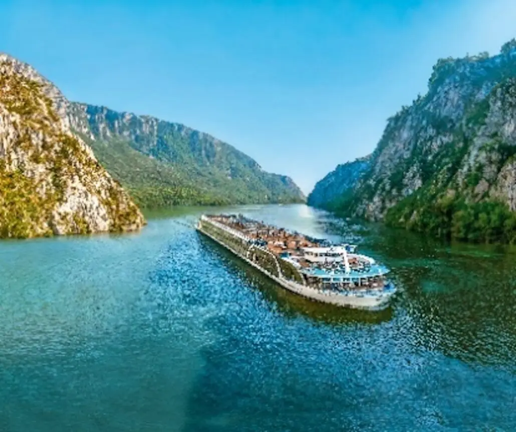 A large river cruise ship travels between steep, green hills on a wide river under a clear blue sky. The water is calm and reflects the landscape.