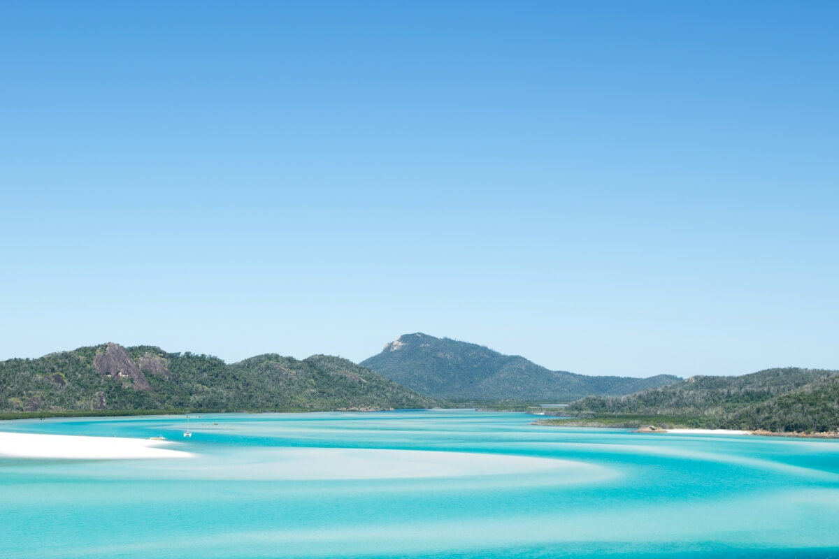 Bright turquoise Australia peaceful waters flow around white sandbars in the foreground, with green hills and a clear blue sky in the background—perfect for discovering quiet corners or enjoying yacht exploration.