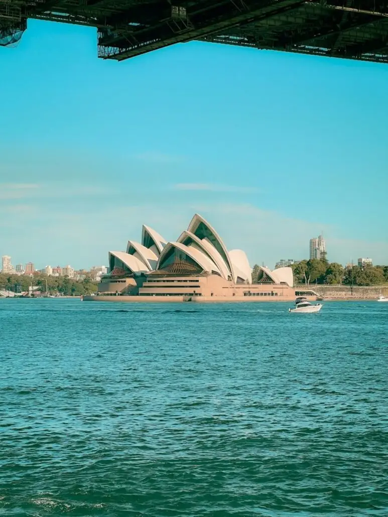 The Sydney Opera House is seen across peaceful waters under a clear blue sky, with an Australia yacht on the harbor and part of a large bridge visible at the top of the image.