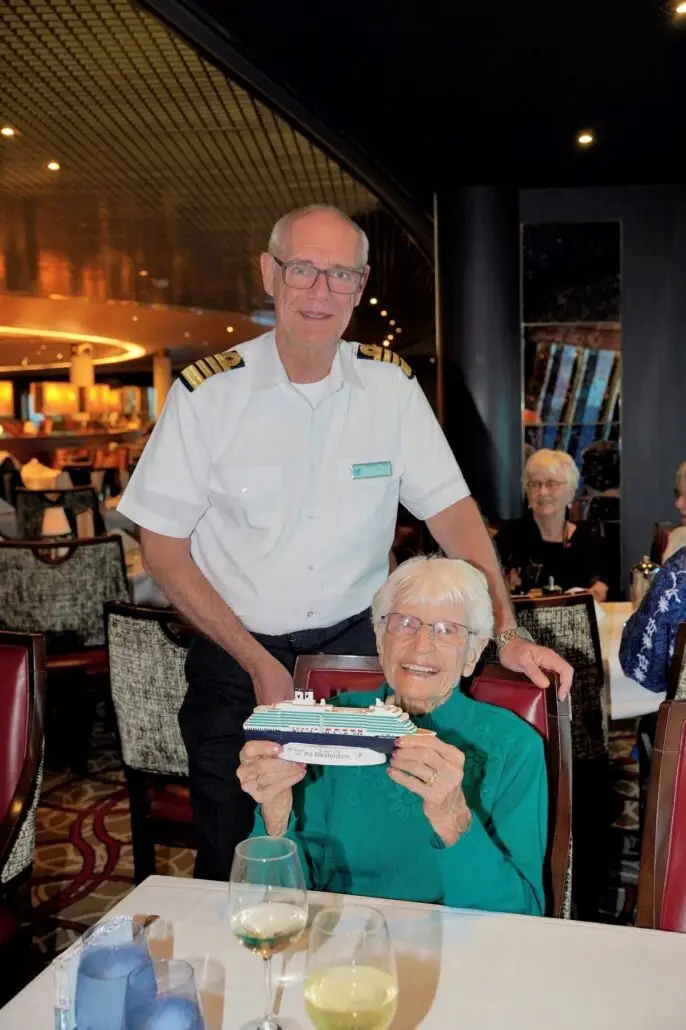 A 106-year-old woman, a former cruise passenger, sits at a dining table holding a model cruise ship, while a man in a captain's uniform stands behind her. Other people are seated at nearby tables in the well-lit restaurant.