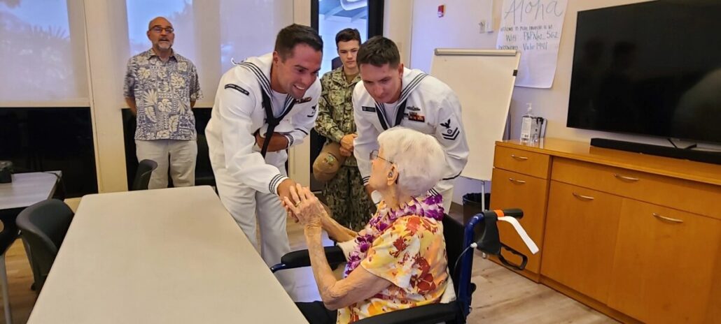 Two sailors in uniform shake hands with a 106-year-old woman in a wheelchair, wearing a flower lei. Two others stand in the background—one in uniform and one in a patterned shirt. The indoor setting hints at a Pearl Harbor tribute.