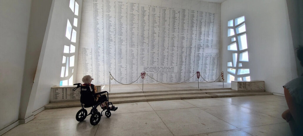 A 106-year-old cruise passenger in a wheelchair gazes at the white marble wall inscribed with names at the USS Arizona Memorial in Pearl Harbor, Hawaii. Natural light pours through geometric windows on the right side.