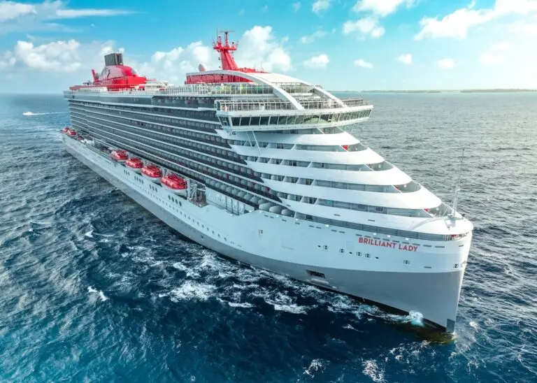 A large white and red cruise ship named "Brilliant Lady" sails on the ocean under a partly cloudy blue sky, with the coastline visible in the distance.