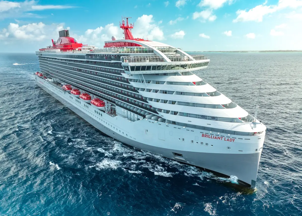A large white and red cruise ship named "Brilliant Lady" sails on the ocean under a partly cloudy blue sky, with the coastline visible in the distance.