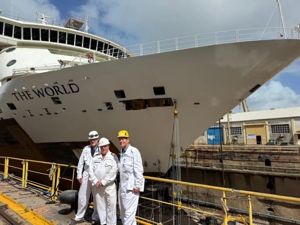 Three people in white coveralls and hard hats stand in front of the World Residential Yacht, docked at a major dry dock. The ship's bow and part of the $30 Million Dry Dock shipyard infrastructure are visible.