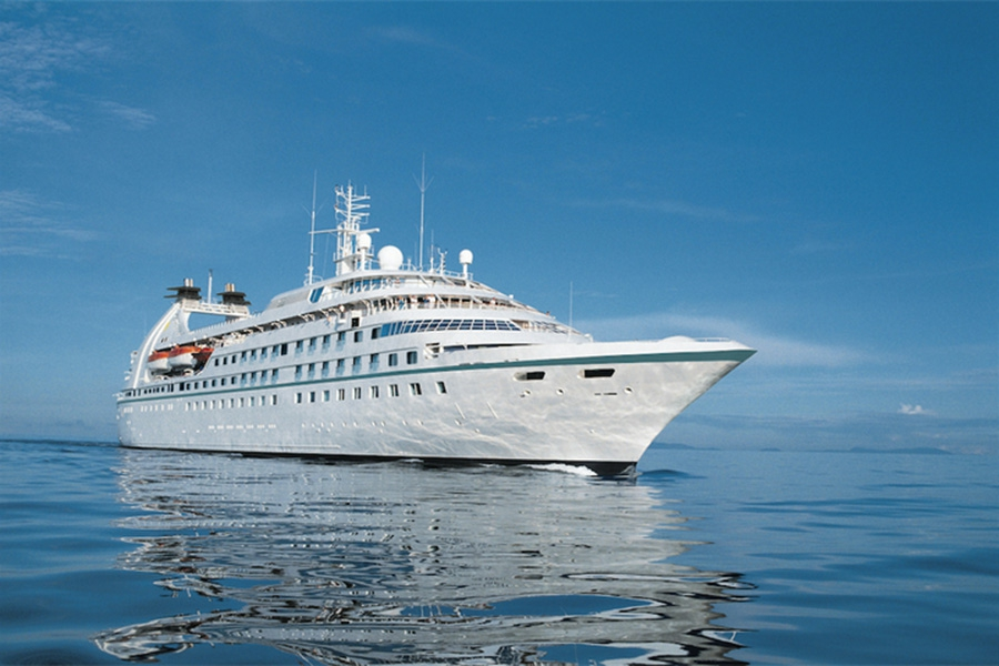 A large white cruise ship is shown floating on calm, reflective blue water under a clear blue sky. The ship has multiple decks and various antennas on top.