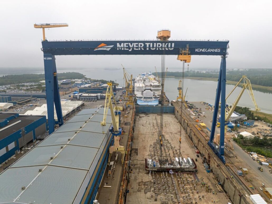 A large shipyard with a blue gantry crane labeled “Meyer Turku” spans over a dry dock. Showcasing advanced shipbuilding, several ships for Royal Caribbean are under construction as cranes and buildings surround the dock on a cloudy day.