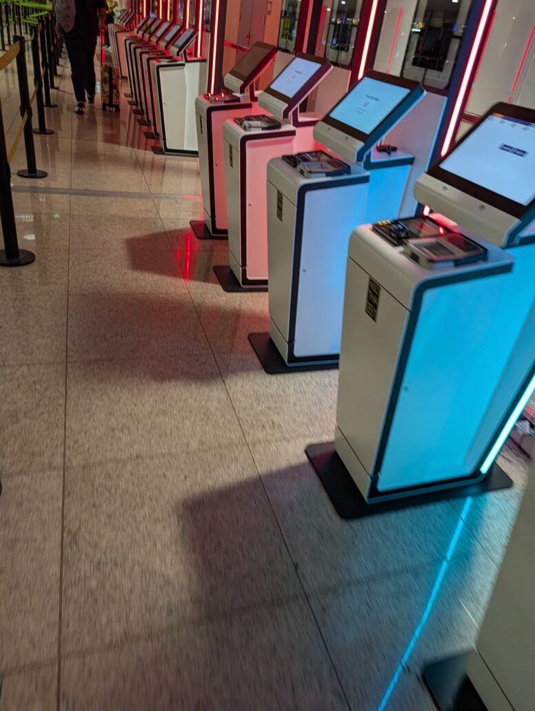 A row of self-service kiosks with screens and passport scanners, illuminated with red and blue lights, is positioned along a tiled floor in what appears to be an airport, welcoming travelers for the Star Princess inaugural cruise.