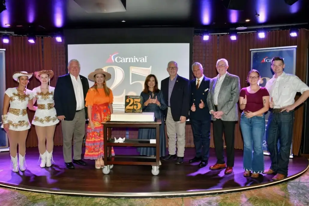 A group of ten people, including four in costumes, pose indoors at the Port of Galveston in front of a Carnival 25 Years anniversary sign and a cake with a gold “25” topper. Some people are smiling and giving thumbs-up gestures.