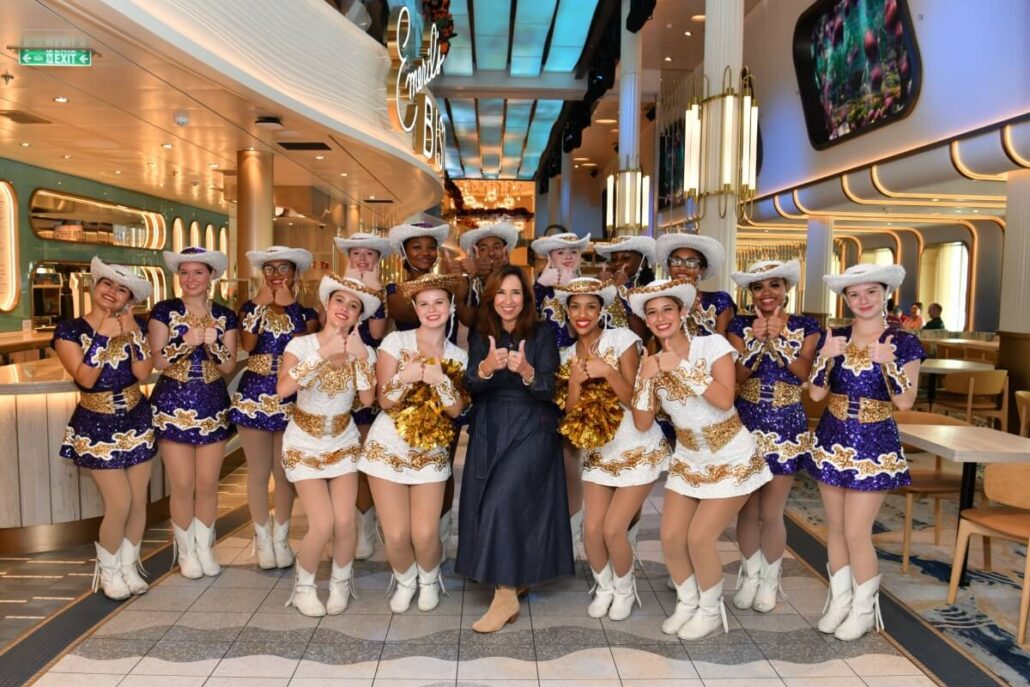A group of dancers in white and gold uniforms with cowboy hats pose with a woman in a dark dress at a brightly lit, modern venue. Decorative lights and tables hint at a festive Carnival event celebrating 25 Years at the Port of Galveston.
