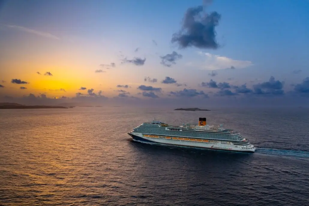 A large cruise ship, the Carnival Venezia, sails across calm ocean waters at sunset near Miami, with an orange and blue sky and scattered clouds. Small islands are visible on the horizon as Carnival expands Florida operations.