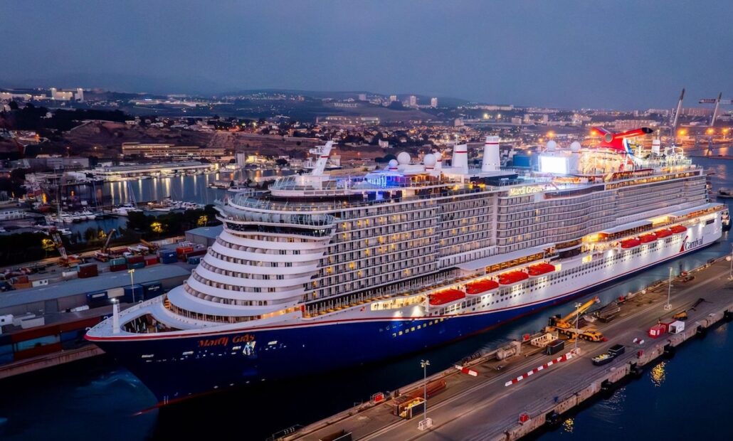 The Carnival Mardi Gras, a large cruise ship, is docked at a port in the evening, brightly lit with multiple decks visible. City lights and buildings form the backdrop as the ship returns to service after dry dock.