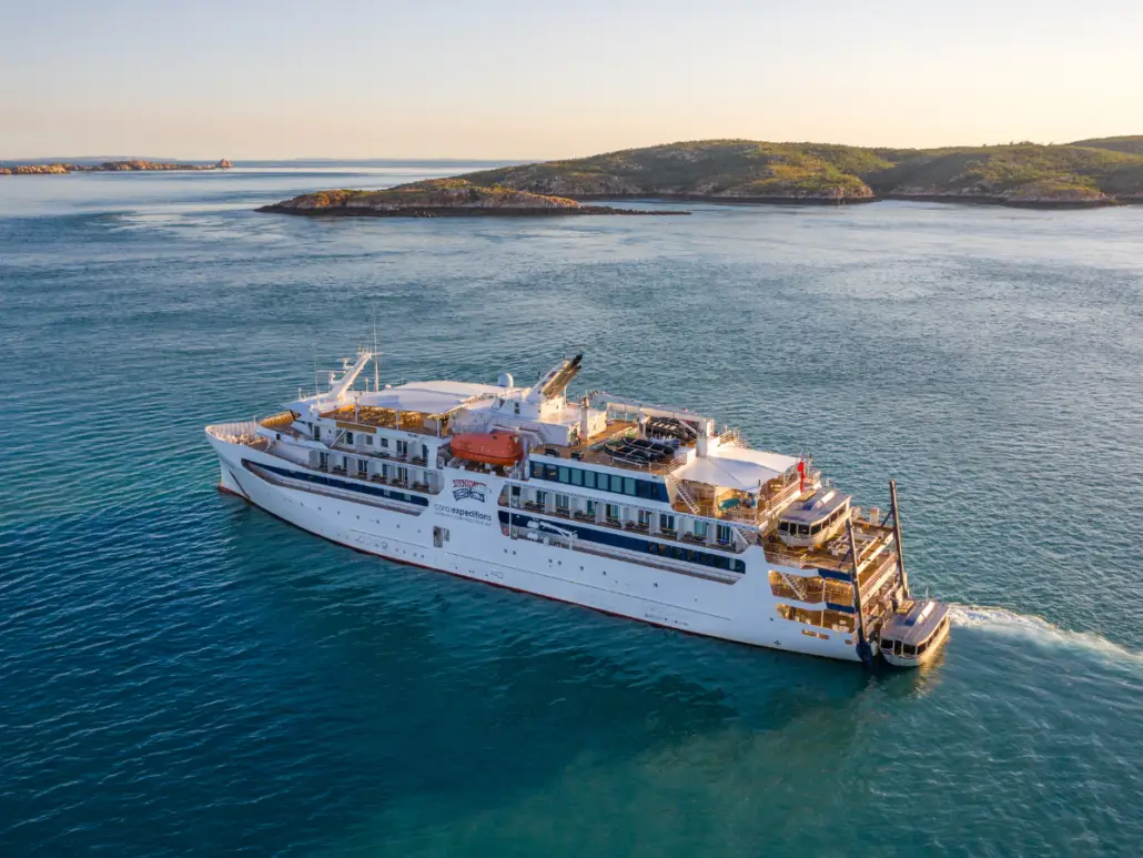 A white cruise ship sails on calm blue water near rocky, green islands under a clear sky. The ship has multiple decks and open areas for passengers, ensuring no Cruise guest is left behind on island adventures.