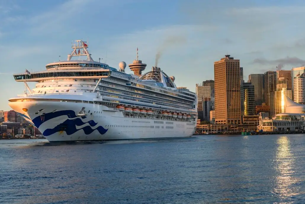 A large white cruise ship is docked at a city port in Vancouver, with modern high-rise buildings and a glass-domed structure in the background—representing the thriving Vancouver cruise industry that generated over $1 billion in 2025. Warm sunrise or sunset light fills the scene.