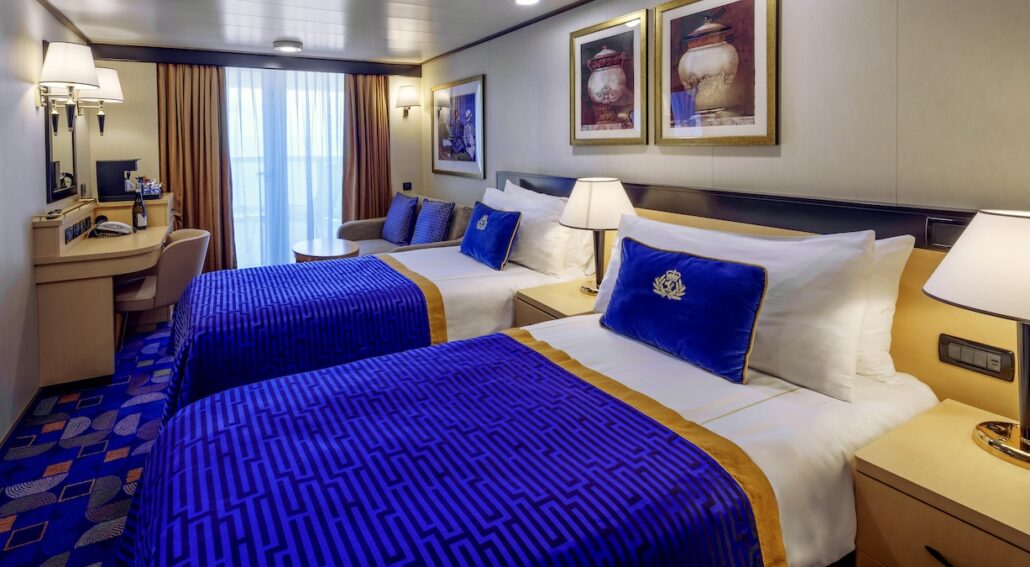 A hotel room with two single beds featuring blue and gold bedding, a desk with a chair, wall-mounted lamps, framed artwork above the beds, and a window with curtains evoking the elegance of Queen Elizabeth during Caribbean Season.