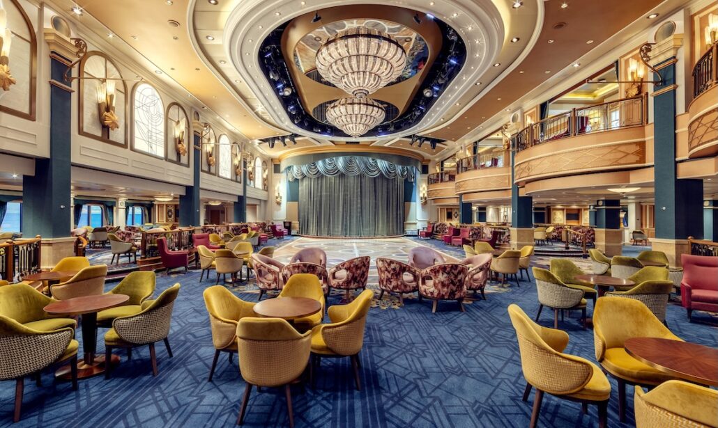 A spacious, elegantly decorated theater lounge aboard Queen Elizabeth features round tables, colorful upholstered chairs, a large stage with a blue curtain, chandeliers, and a high ceiling—perfect for Caribbean Season departures from PortMiami.