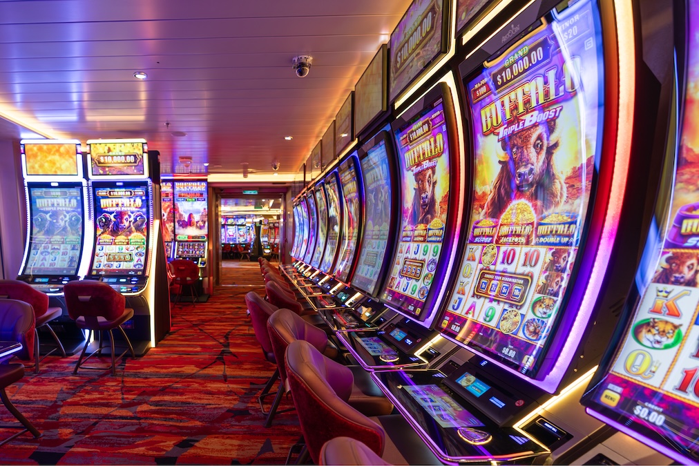 A row of brightly lit slot machines lines one side of the Carnival Mardi Gras casino, featuring colorful carpeting and empty chairs. The environment is clean and modern, recently refreshed after dry dock, with ceiling lights and digital displays.