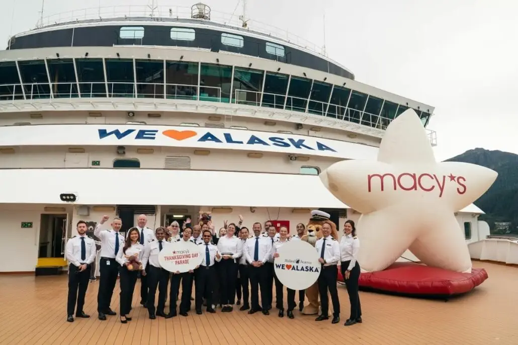 A group in uniform stands on the deck of a Holland America Line ship with "WE ♥ ALASKA" and a large Macy's star balloon displayed behind them, celebrating their Alaska Float for the Macy's Thanksgiving Day Parade. The bridge and a mountain are also visible.