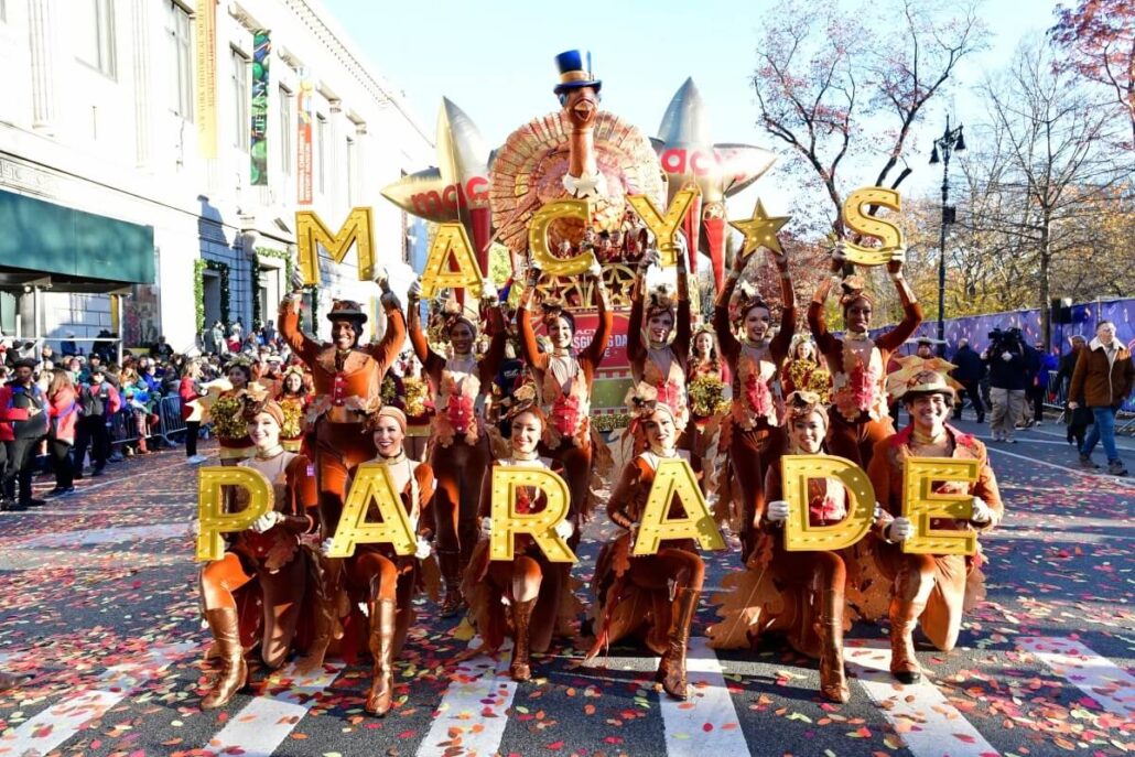 A group of performers in orange costumes pose on a street holding large gold letters that spell "MACY'S PARADE" during the Macy's Thanksgiving Day Parade. A large turkey float and spectators are visible, with confetti covering the ground.
