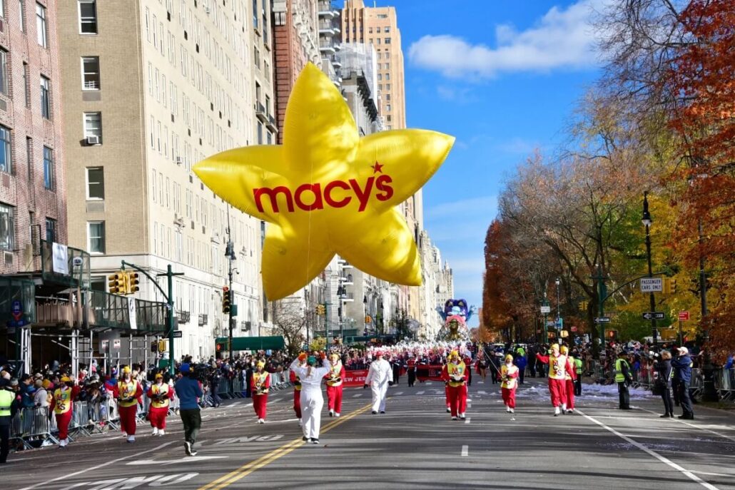 People walk down a city street guiding a large yellow star-shaped Macy's balloon during the Macy's Thanksgiving Day Parade. Spectators line the sidewalks, while autumn trees and tall buildings create a festive backdrop.