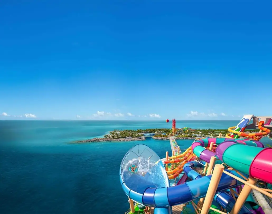 A person slides down a clear, twisting water slide surrounded by colorful slides on Royal Caribbean’s Legend of the Seas, with an island and blue ocean in the background under a clear sky.