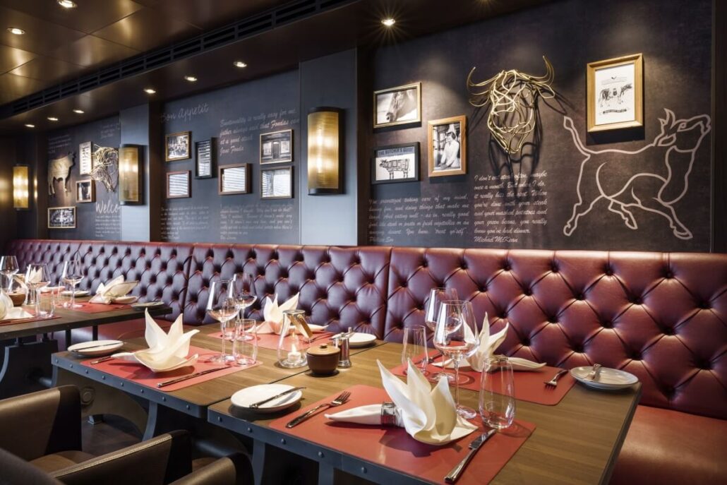 A modern restaurant dining area in Galveston featuring red tufted leather seating, set tables with folded napkins and wine glasses, chalkboard-style wall art, framed photos, and ambient lighting—perfect for savoring Texas-inspired flavors.