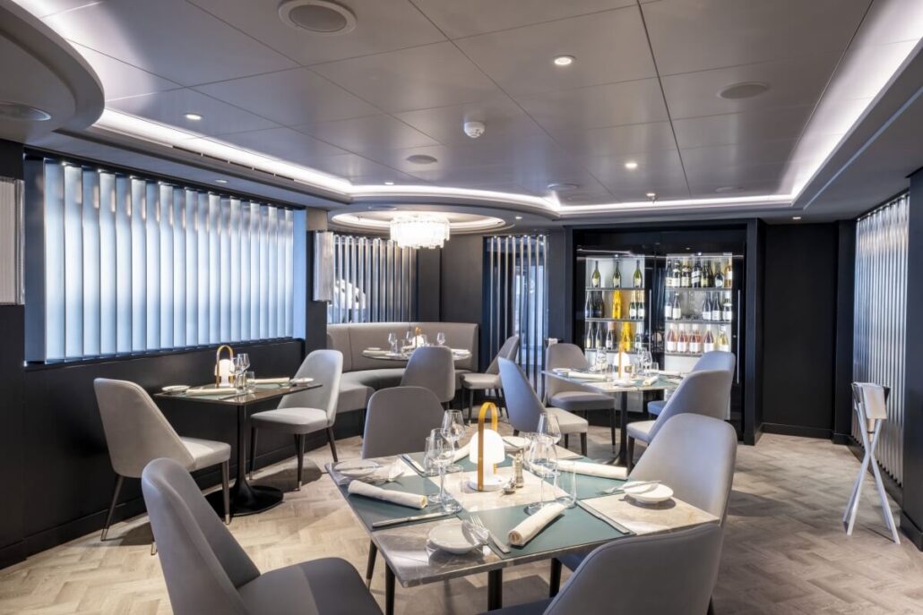 Modern restaurant dining area on MSC Seascape, featuring gray chairs, square tables set with glasses and napkins, a chandelier, large windows with vertical blinds, and a glass wine display case—perfect for savoring Texas-inspired flavors.