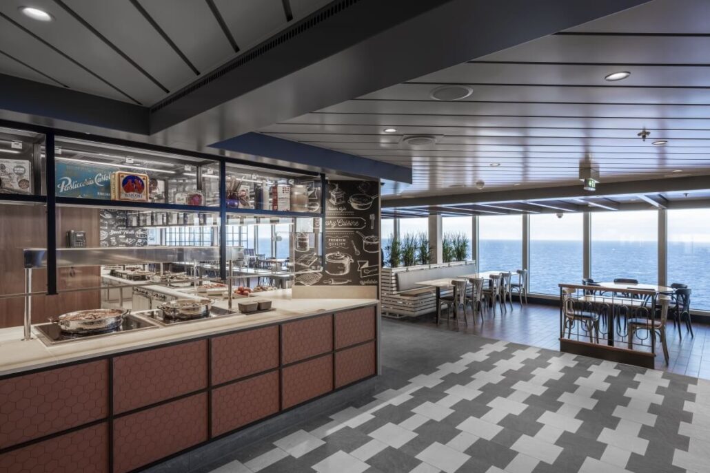 A modern self-service cafeteria on MSC Seascape with food stations, light wood tables and chairs, large windows overlooking the ocean, a patterned tile floor, and wall art featuring Texas-inspired flavors and vibrant food illustrations.
