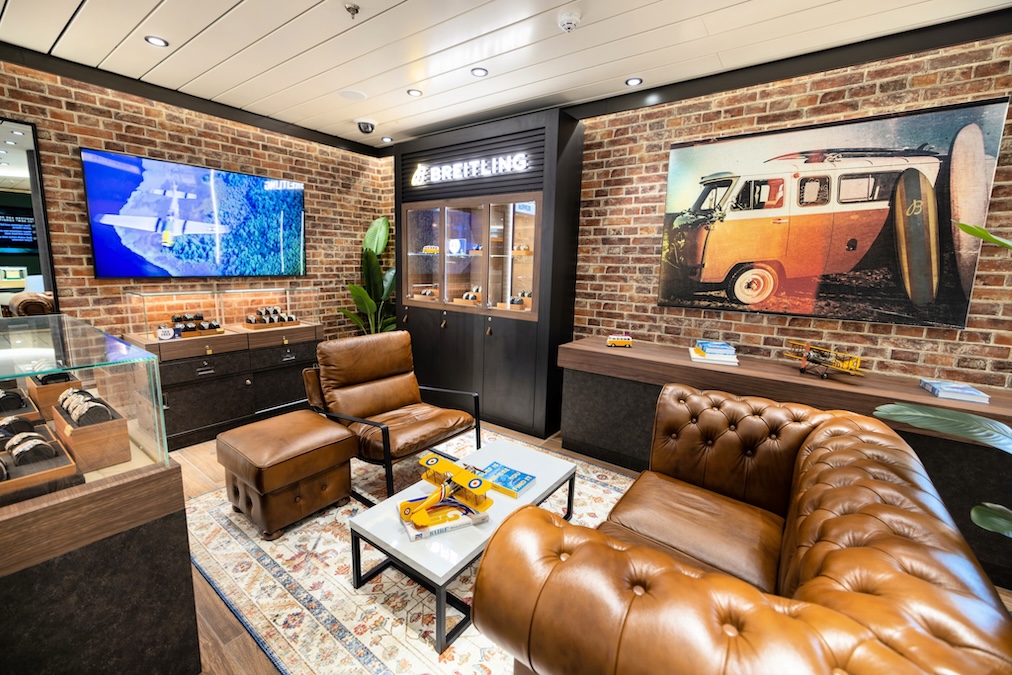 A modern store lounge with brown leather sofas and chairs, a display case of watches, a model yellow airplane on the coffee table, brick walls, a Breitling sign, and a large photo of Carnival Mardi Gras returning to service with a surfboard-topped vintage van.