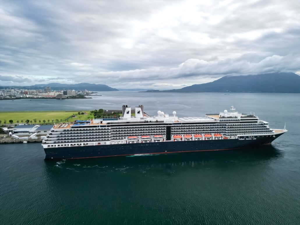 A large Holland America cruise ship is docked near a green shoreline, with a city visible in the background and mountains across the water under a cloudy sky, showcasing the allure of Asia cruises and scenic Japan ports.