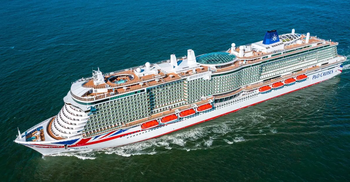 A large white P&O Cruises ship, possibly Iona, with multiple decks and orange lifeboats sails on calm blue water. The vessel features a glass-covered upper deck area and the “P&O Cruises” logo is visible on its side.