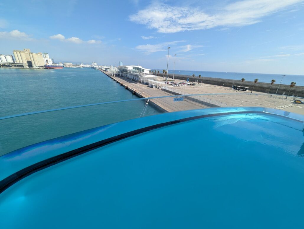 Enjoy a relaxing view from the pool aboard the Star Princess, overlooking a port with palm trees, industrial buildings, and a dock—all under a partly cloudy sky.