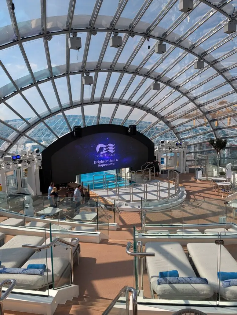 A glass-domed indoor lounge on the Star Princess features padded lounge chairs, a stage with steps, and a large digital screen showing the Princess Cruises logo. Two people stand near the stage, perhaps ready for an evening of dining or to share a trip report.