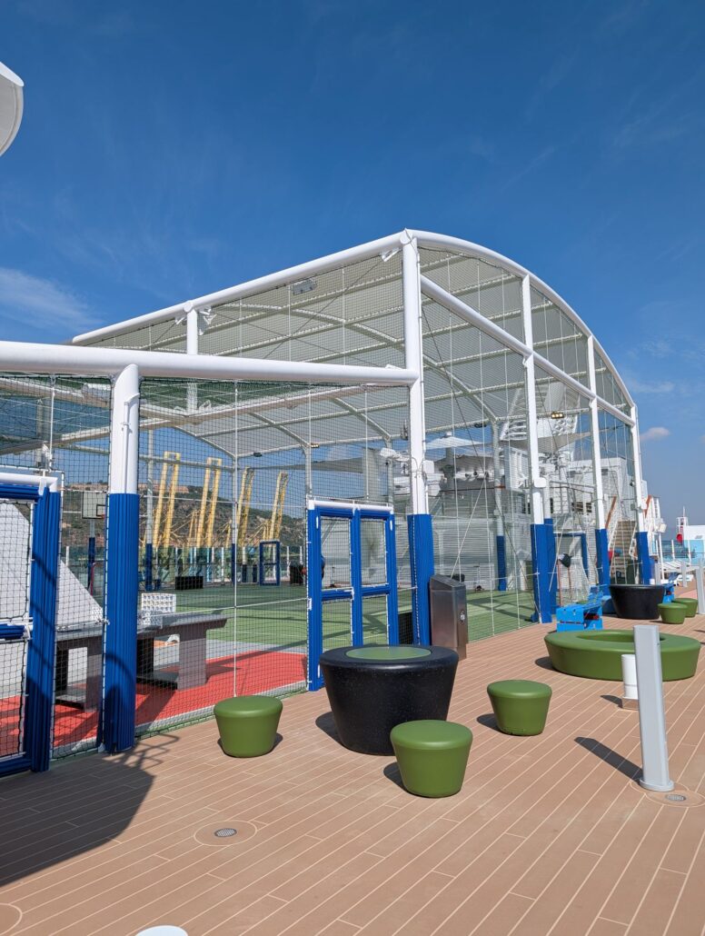 Outdoor sports court on the Star Princess cruise ship, surrounded by a tall netted enclosure. Green seating and a table for dining are in the foreground on a wooden deck, with clear blue sky overhead.