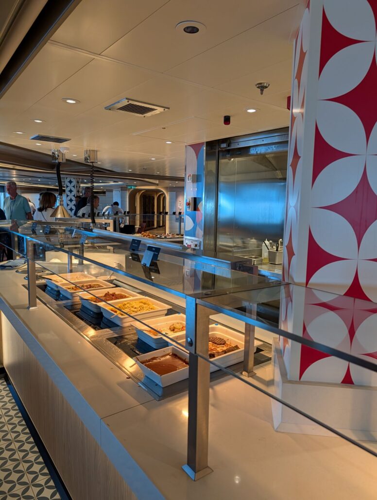 A self-service buffet dining area with trays of hot food on a counter behind glass, with a few people in the background selecting items. The area is clean and brightly lit, featuring geometric red and white wall decor.