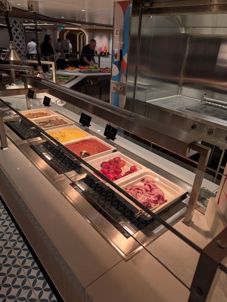 A buffet counter on the Star Princess offering trays of bacon, scrambled eggs, hash browns, baked beans, grilled tomatoes, and sliced ham. Dining labels sit above each tray as people in the background add to their trip report experience.