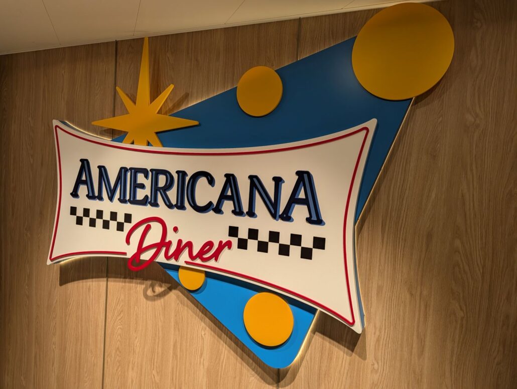 A sign reading "Americana Diner" in bold letters with a retro design, featuring blue and yellow geometric shapes and a starburst, mounted on a wooden wall—a picture-perfect spot for your next dining trip report.
