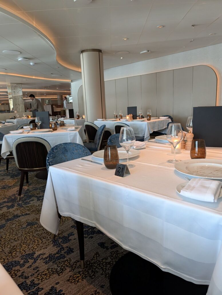 A modern dining restaurant with tables set for dinner, featuring white tablecloths, brown glassware, and elegant plates. Candles and black menu covers accent some tables. A person can be seen in the background near the kitchen—a scene fit for a Star Princess trip report.