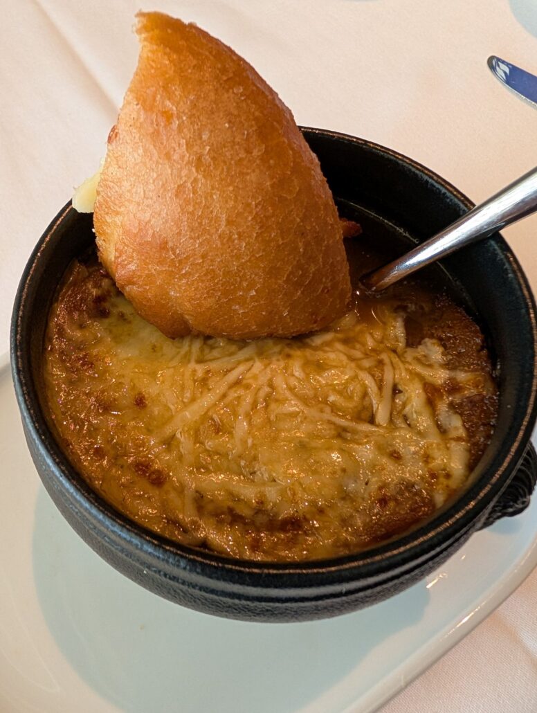 A small black bowl of French onion soup topped with melted cheese and shredded cheese, served with a slice of toasted bread and a metal spoon—just one highlight from the Star Princess dining experience featured in my trip report.