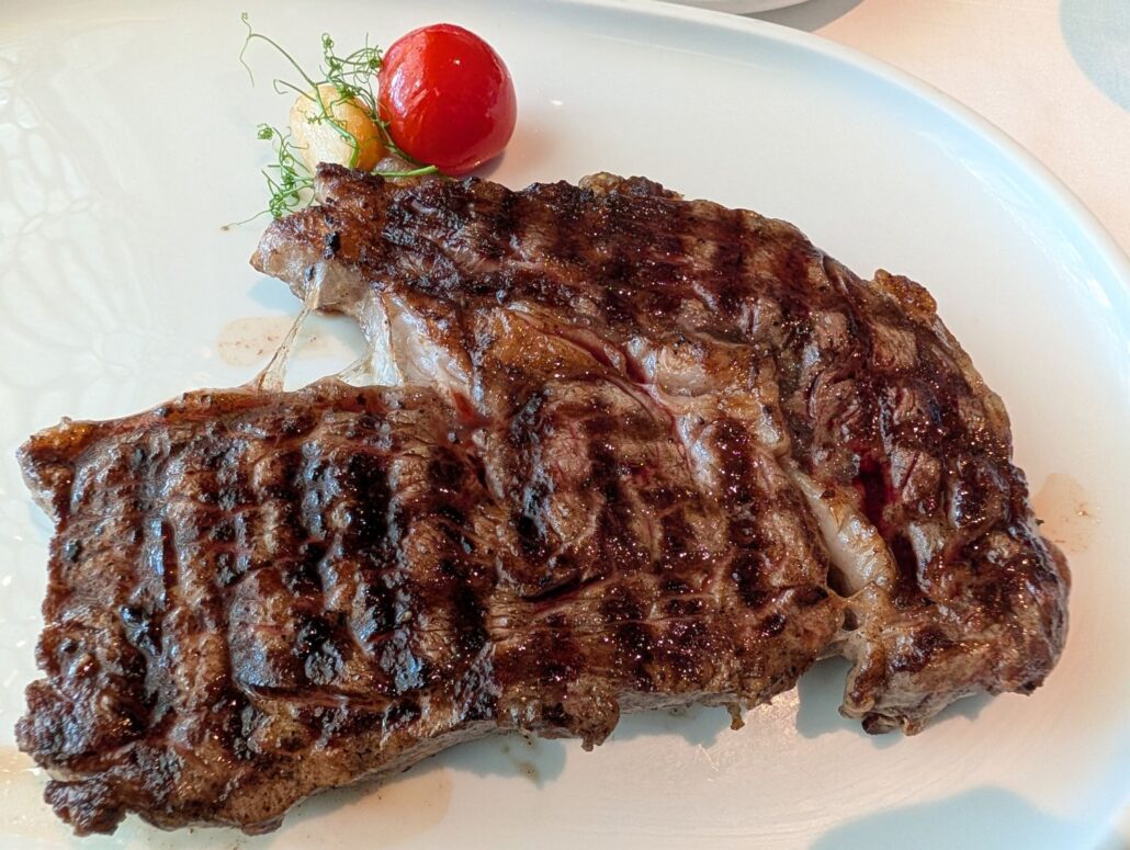 A grilled steak with visible char marks is served on a white plate, accompanied by a small tomato and a garnish of herbs—a delicious highlight from the Star Princess dining experience mentioned in our trip report.