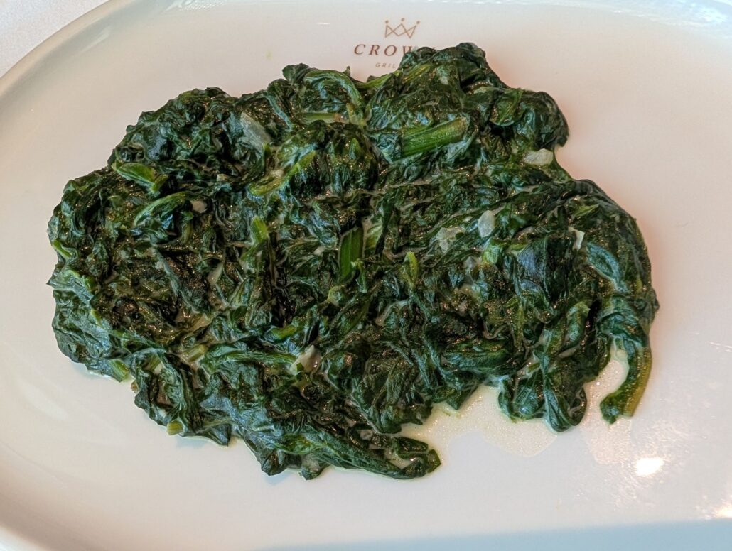 A serving of cooked spinach is spread out on a white plate, with a small amount of visible moisture or oil. The plate, featuring a faint gold logo and text near the top edge, adds an elegant touch to the Star Princess dining experience.