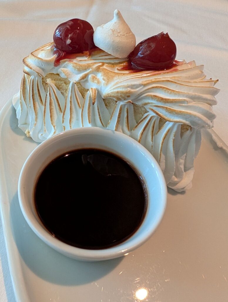 A slice of cake topped with toasted meringue, two cherries, and a small meringue, served on a white plate with a small dish of dark sauce—an elegant treat highlighting Star Princess dining.