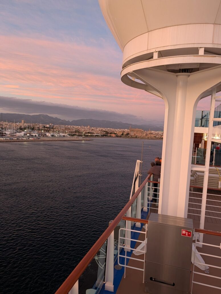 View from the deck of the Star Princess at sunset, showing two people seated along the railing, calm water below, a distant city and marina, and a partly cloudy sky with pink and purple hues—perfect for a trip report or recalling memorable dining moments.
