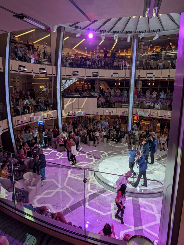 A large crowd sits and stands around a circular stage in an indoor atrium with multiple balconies. Several people dine and mingle on the stage under purple lights, while others watch from various levels—perfect for a Star Princess trip report.
