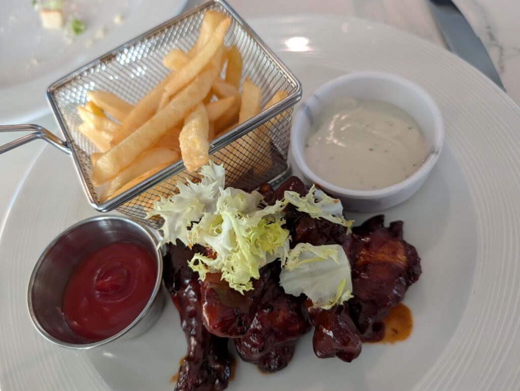 A white plate with French fries in a metal basket, a small cup of white dipping sauce, ketchup, and BBQ-glazed meat pieces topped with green lettuce—perfect for savoring during your Star Princess dining experience or sharing in a trip report.